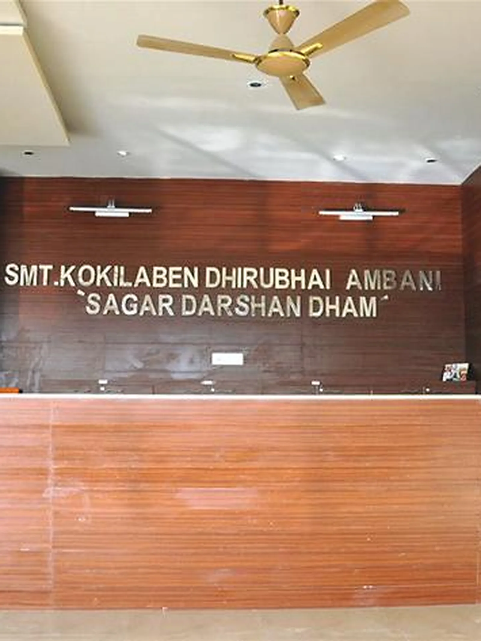 Reception desk with a dedication board behind it reading Smt. Kokilaben Dhirubhai Ambani, Sagar Darshan Dham